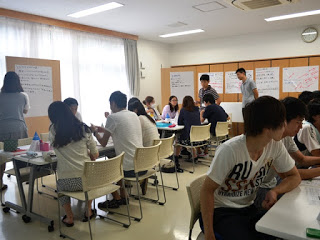 明るい教室内で複数のグループがそれぞれテーブルを囲んでボードゲームに取り組んでおり、壁には模造紙に書かれた発表資料が貼られている様子の写真