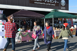 「南三陸ポータルセンター」と書かれた建物の前で、黒い屋根の屋台と緑の屋根の屋台があり、それぞれのテーブルには装飾品や食べ物と思われる品が並べられ、左側には大きな雪だるまの飾りが見え、人々が屋台の前を歩いている屋外イベントの写真