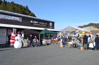 南三陸ポータルセンターの建物の入り口前に、風船などで飾り付けられた複数のテントが並び、傍らにサンタクロースと雪だるまの大型飾りが飾られ、たくさんの来場者で賑わっているクリスマス市の様子の写真