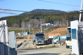 山と森林、田畑のそばにある建設現場で、黒い大型トラックや青い小型トラック停車しており、工事用のバリケードや壁が立っている様子の写真