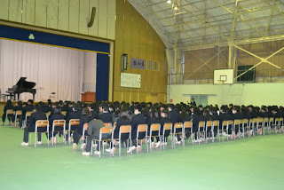 ステージ上中央にはグランドピアノがあり、右側にはバスケットゴールも見える広い体育館の中に、ステージの方を向いた制服を着た生徒たちが椅子に整然と座っている後ろ姿の写真