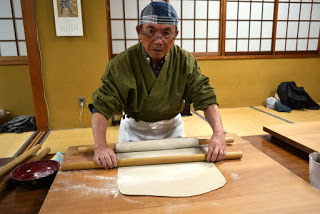 畳の床と障子がある和風の部屋で、緑色の作務衣の上着にエプロンと頭布を身につけた男性が、大きな麺棒で木の台の上の生地を伸ばしている作業中の写真