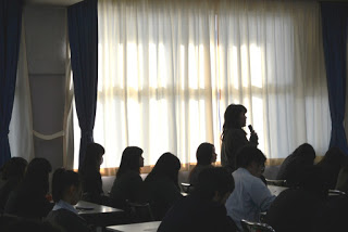 カーテン越しの光が差し込む薄暗い教室で、窓際に立つ女子生徒が町長に質問を行っている様子を、他の学生たちが机に座って話を聞いている写真