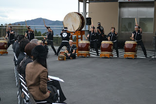 屋外で、お揃いの衣装を着た複数の人々が大小さまざまな和太鼓を演奏しており、手前には椅子に座って鑑賞する来賓の方々、奥には建物と遠くに山々が見えるおおもり創作太鼓の演奏の写真