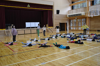 体育館で、子供たちが床に仰向けに寝転び、周囲に数人の大人が立って見守っている様子が写っている写真