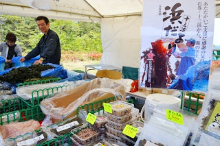 白いテントの下で、容器やパッケージに入った様々な海産物が黄色い値札とともに並び、奥には「浜だより」と書かれた大きなポスターが飾られている写真