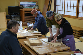 障子や木製家具が設置された和風の室内で木のテーブルを囲み、一人の男性が作業に集中し、向かい側で他の男性が見ているなか、男性講師に教わりながら紫色のエプロンを着用した参加者の女性が、粉をまぶして伸ばした蕎麦を包丁で切っているそば打ち体験の様子の写真