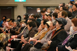 ピンク色のプログラムを手に持ち、ホールの観客席に座った高齢の人々が舞台を見ながら笑顔で催しを楽しんでいる様子を写した写真