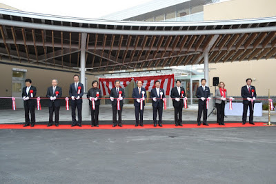 屋外の完成した病院の前で、赤いカーペットの上で正装した11人の人物が並び、それぞれが赤いリボンをハサミで切っているテープカットの様子の写真