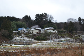 雪が部分的に積もった斜面にいくつかの家が建ち、背景には木々が広がり、曇り空の下で冬の雰囲気が漂う風景の写真