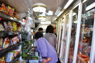左側にスナックやインスタント食品などの食品が並ぶ棚があり、右側には肉類などの生鮮食品が入った冷蔵ケースが設置された、狭いながらも整理された明るい移動販売車内で数人の客が商品を選んでいる様子が写っている写真