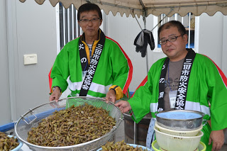 男性二人が緑の法被を着て大きなザルに入った大量の煮豆のような料理を持ち、テントの下で並んでいる写真