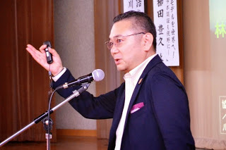 紺色のスーツを着た男性がマイクの前でプレゼン用のリモコンを手に話している講演中の登壇者の写真