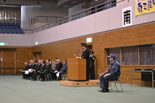 ベイサイドアリーナの屋内会場で、壇上に立つ町長が出初式の挨拶を行い、その横に制服姿の関係者が控え、後方には来賓と見られる多くの人が椅子に座って整然と並んでいる出初式の写真