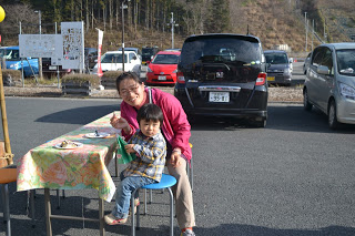 屋外のテーブル席に座っているピンクのジャケットを着た女性と、チェック柄のシャツを着た男の子の親子が笑顔でカメラを見ている写真