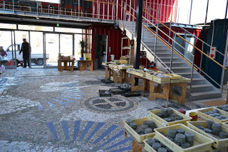 モザイク模様の床が広がる屋内空間に、さまざまな物が置かれたテーブルと上階へ続く階段があり、円柱状の物体が並ぶトレイが複数置かれた自然光が差し込む明るい室内の写真