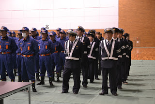 べイサイドアリーナの屋内会場で、青い制服の消防団員と黒い制服に白いたすきを掛けた交通指導隊員が、列を作って厳粛な面持ちで立っている出初式の写真