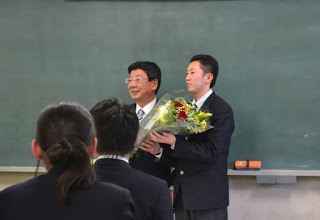 教室の前方で、佐藤町長と男子生徒が一緒に花束を持って並んで立ち、他の生徒たちが座ってその様子を見守っている写真