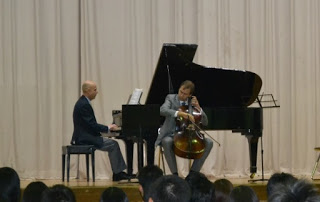 手前で生徒たちが見ているなか、背景にはベージュ色のカーテンがひかれたステージ上で2人の演奏者が演奏しており、1人はグランドピアノを弾き、もう1人はチェロを演奏している写真