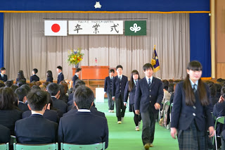 「卒業式」と書かれた横断幕と校旗、壇上の演台や花が飾られたステージ下で、両側に座る生徒たちの間の通路を制服姿の卒業生が通路を歩いている、厳かな式典の写真