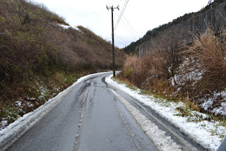 曇り空の下、雪がシャーベット状に残る山道が右にカーブし、濡れた路面が光る冬の風景