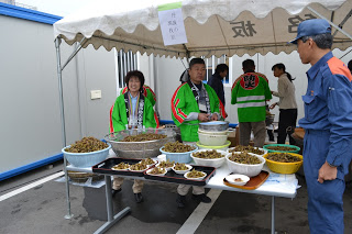 テントの下で緑の法被を着た人たちが、大量の煮豆などを皿に盛って配膳の準備をしている写真