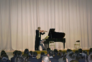 手前で生徒たちが演奏を見ているなか、背景には大きなカーテンが広がるステージ上でピアノ奏者がグランドピアノに座って演奏しており、前でバイオリン奏者が立って演奏している写真