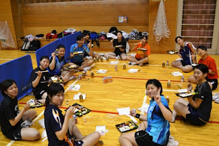 体育館の床に円になって座り、ユニフォーム姿の中学生たちが弁当を食べながら笑顔でピースサインをしている写真