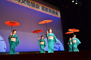 緑色の着物を着た女性たちが赤い和傘を手に持ち、ステージで並んで踊っている写真