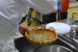 キッチンで、料理人と思われる人物が竹製のざるに入った茹でた麺を白い皿に移している様子が写っている写真