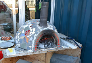 モザイク模様の外装を持つ薪窯が木製のテーブルの上に設置され、周囲には道具や物が置かれ、左側にはトレイに乗ったピザがあり、上部には煙突があり、内部には赤く燃える薪が見えるピザ窯の写真