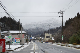 一直線に伸びている道路の左側の歩道に雪が積もったコカ・コーラの自動販売機や看板が並ぶ建物があり、奥には山々が広がる冬の風景の写真