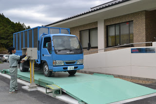 水色の計量器の上に青いトラックが乗り、作業服を着た人が横で操作している写真