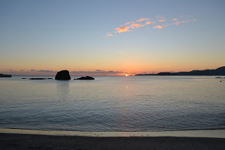 地平線から浮かんできた太陽が空と海を温かく照らし、雲が点在する中、岩が海に浮かび、滑らかな砂浜が緩やかに湾曲して続く朝の静かな海岸の写真