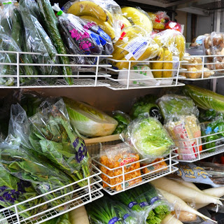 白い網棚に、バナナ、きゅうり、きのこ、葉物野菜などの新鮮な野菜や果物が並べられ、多くがビニール袋に包まれて陳列されている様子が写っている写真