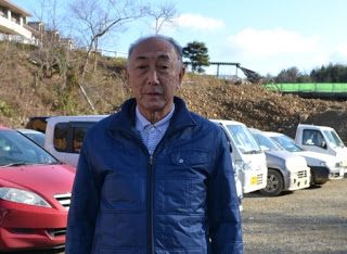 敷地内に複数の車が駐車している前で、青いジャケットと襟付きシャツを着た阿部一郎さんが立っている写真