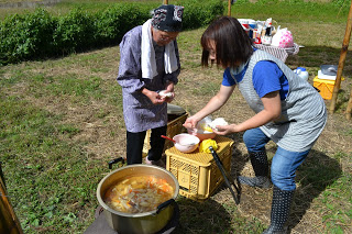 畑のそばでエプロン姿の女性2人が、鍋の煮物を紙の器に盛り付けて昼食の準備をしている写真
