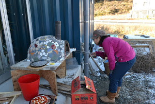 屋外で石造りのモザイクタイルが施されたピザ窯を使い、ピンクのジャケットと青いジーンズを着た女性が窯の火を調整しており、近くには赤いバケツ、ピザピール、薪の入った開いた箱が置かれているピザ窯作業中の写真