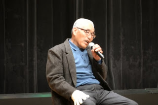 黒いカーテンの前のステージ上で、マイクを持って椅子に座り語っている白髪の高齢男性が、話をしている場面の写真