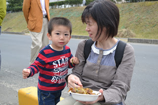 母親が隣に立つ子どもに、煮豆のような料理を入れた器を持ちながら一緒に食べている写真
