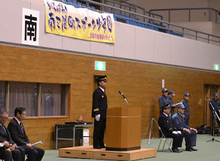 演台の上に横断幕が掲げられ、壁には「南」と書かれた標識があり、周囲には来賓者と制服姿の隊員が座っていたり立っていたりしている会場で、演台の前に立っている制服姿の町長の写真