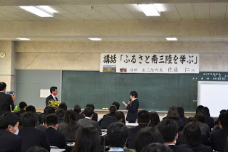 教室の前方で花束を受け取った佐藤町長が「講話『ふるさと南三陸を学ぶ』」と書かれた横断幕の下、マイクを持って話をする男子生徒と向かい合って立ち、多くの学生が座ってその様子を見守っている写真