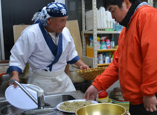流し台で青い頭布をつけた白い服の人が丸皿とざるを持って料理を準備しており、オレンジ色のジャケットを着た別の人が茹で上がった蕎麦が盛り付けられた皿に手を伸ばしている様子が写っている写真