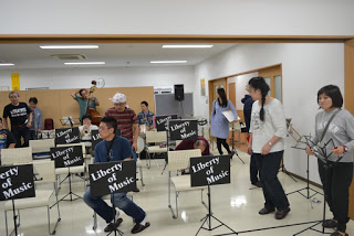 複数の演奏者や関係者がステージ前に集まって準備をしており、「Liberty of Music」の譜面台が並ぶ中で和やかに打ち合わせをしているリハーサル中の写真