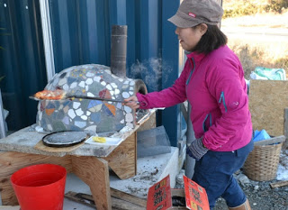 周囲には赤いバケツや木の板、その他の道具が置かれている屋外のモザイク模様のピザ窯のそばに立つ女性が、焼けたばかりのピザを乗せたピザピールを持っている写真