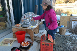 屋外でピンクのジャケットと帽子を身に着けた女性がピザピールに乗せたピザを窯に入れようとしており、周囲には赤いバケツ、トレイに乗った別のピザ、オレンジ色の箱、建築資材や道具が見えるピザ窯作業中の写真