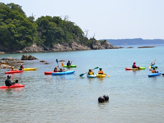 穏やかな海の上に、参加者たちが乗った赤や黄色、水色のカヤックがたくさん浮かんでいる写真
