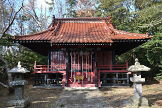 2つの石灯籠の間に石畳の通路があり、その奥に、木々を背景に朱色の社殿が建っている写真