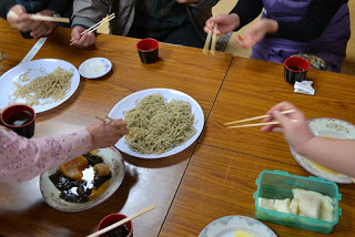 周囲にはさまざまな料理がのった小皿や赤い小さなカップが並び、中央には大皿に盛られた麺料理が置かれた木のテーブルを囲み、数人が打ち立ての蕎麦を実食しようと箸を持った手を伸ばしている様子の写真