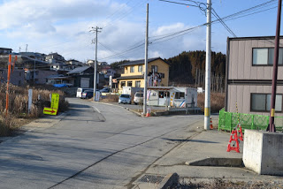 空はところどころ雲があり、背景には木々のある丘と点在する住宅が見え、二股に分かれた左側にカーブした道路が小さな住宅街へと続く郊外の街並みで、中央には黄色い家、右側には灰色の建物と工事用のバリケードや注意喚起の看板が設置されている写真